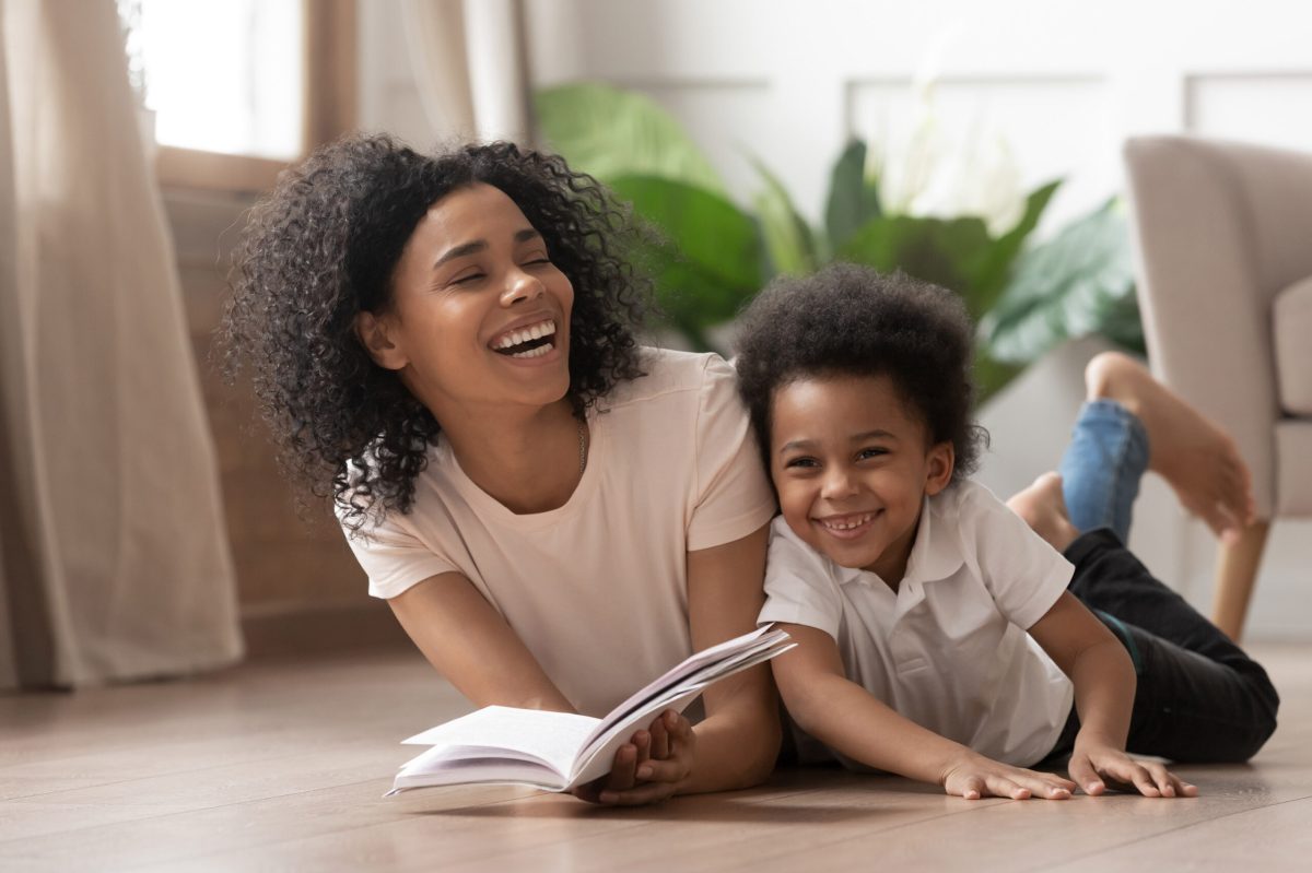 Caring african american mother baby sitter tell funny fairy tale story to smart cute kid son laugh lay on warm floor together, loving mixed race mom read book having fun with little child boy at home